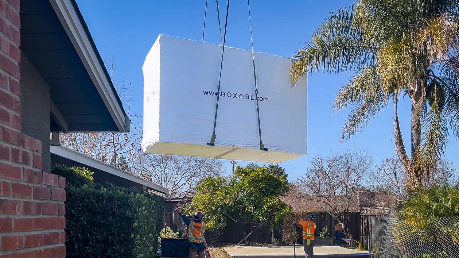 A crane lowering a folded Boxabl Casita over a fence into a Southern California backyard with palm trees